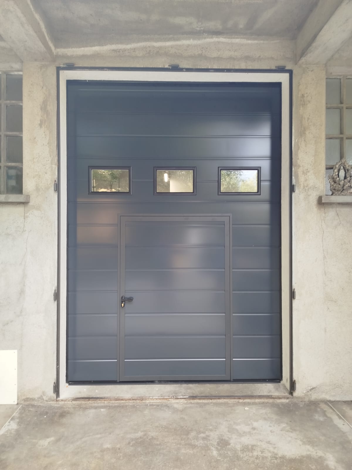 porta sezionale a <p>porta sezionale oltre luce, con portina inserita, oblò e pannello con doga da 25cm. RAL 7016</p>