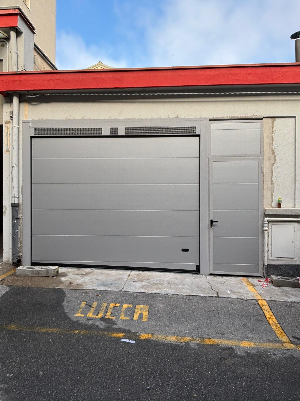 porta sezionale a <p>porta sezionale in luce, con veletta areata, maniglia di sollevamento, coprifili, lampeggiante e pannello con doga da 25cm e portina d'ingresso a lato. RAL 9006</p>