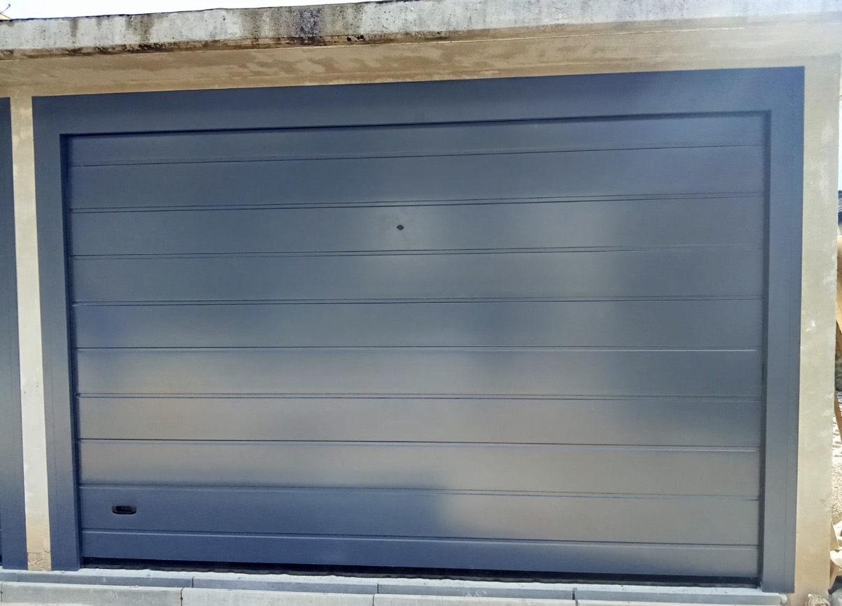 porta sezionale a <p>porta sezionale in luce, con maniglia di sollevamento, sblocco esterno e pannello con doga da 25cm. RAL 7016</p>