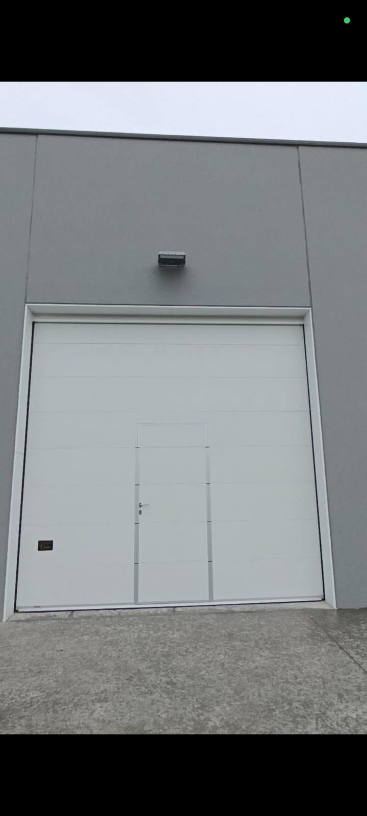 Porta sezionale industriale a <p>Porta sezionale industriale, con portina inserita, maniglia di sollevamento e pannello intero. RAL 9010</p>